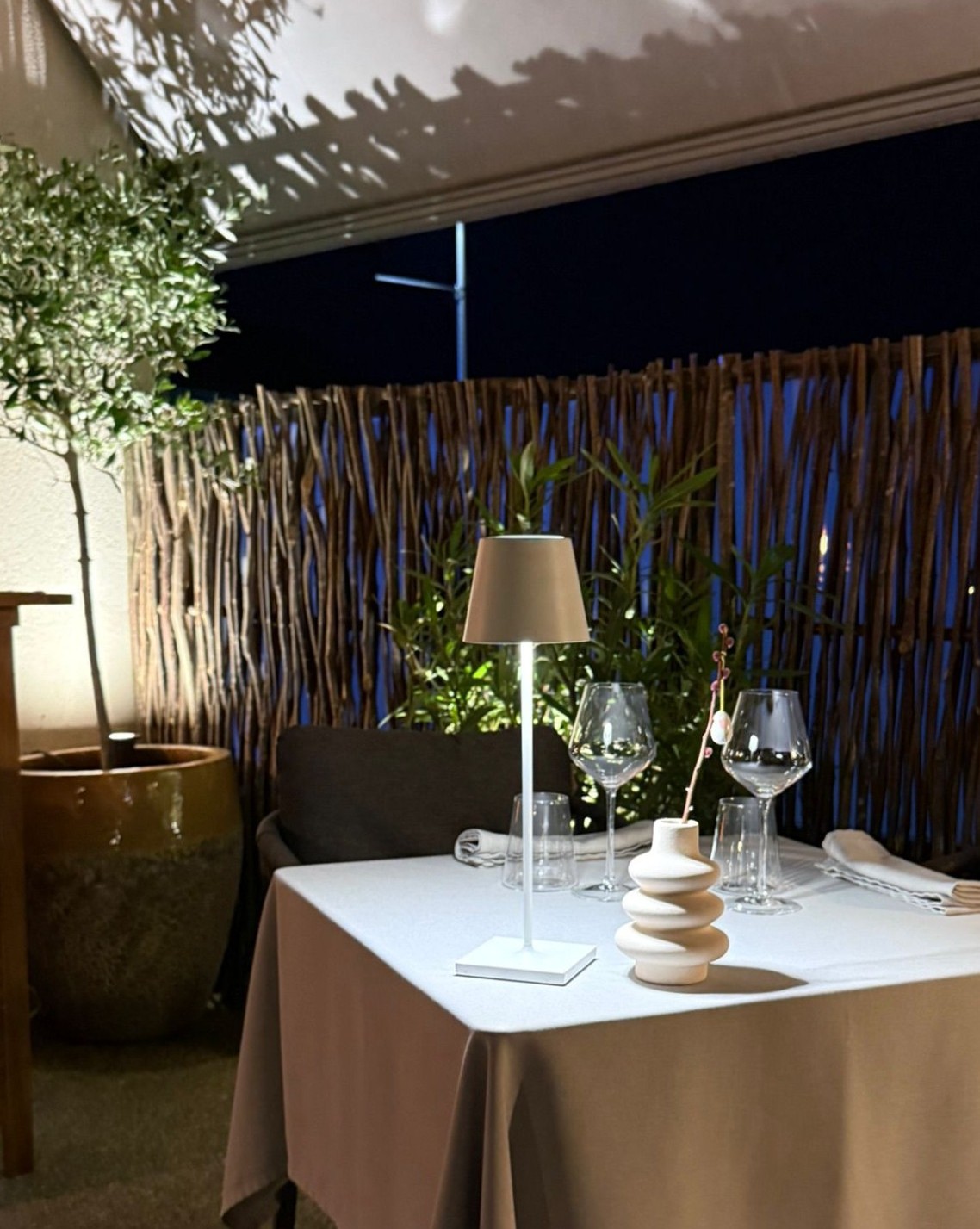 Table pour deux sur la terrasse de l'Auberge d'Arthaz en soirée, nappe taupe, lampe Poldina et vase galets, clôture en branches